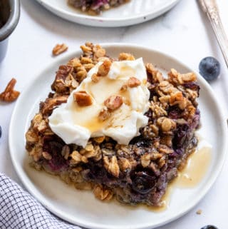 Slice of blueberry baked oatmeal on white plate with maple syrup.