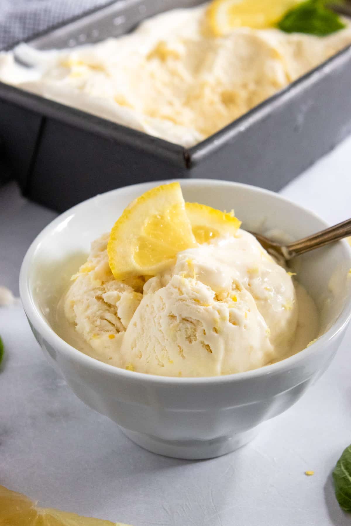 Lemon ice cream in white bowl with lemon wedges and ice cream container behind it.