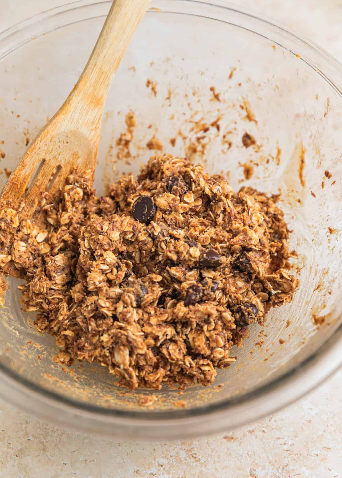 Chocolate chips stirred into oat mixture in glass bowl with wooden spoon.