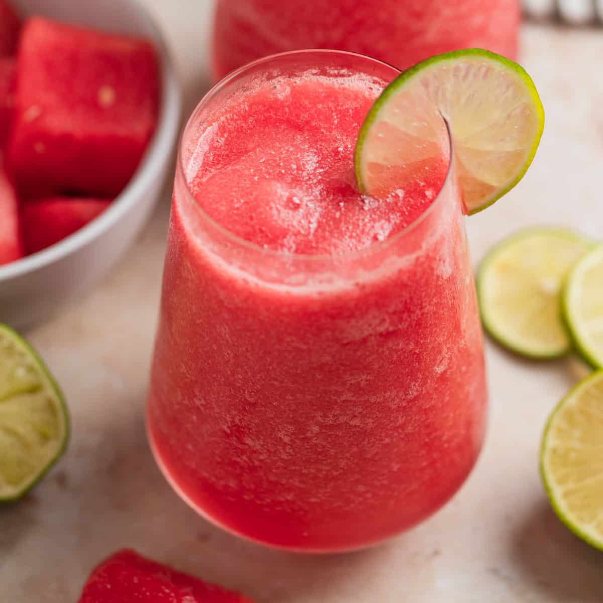 Stemless wine glass with watermelon rosé slushie or frosé with bowl of watermelon behind it as well as lime slices.