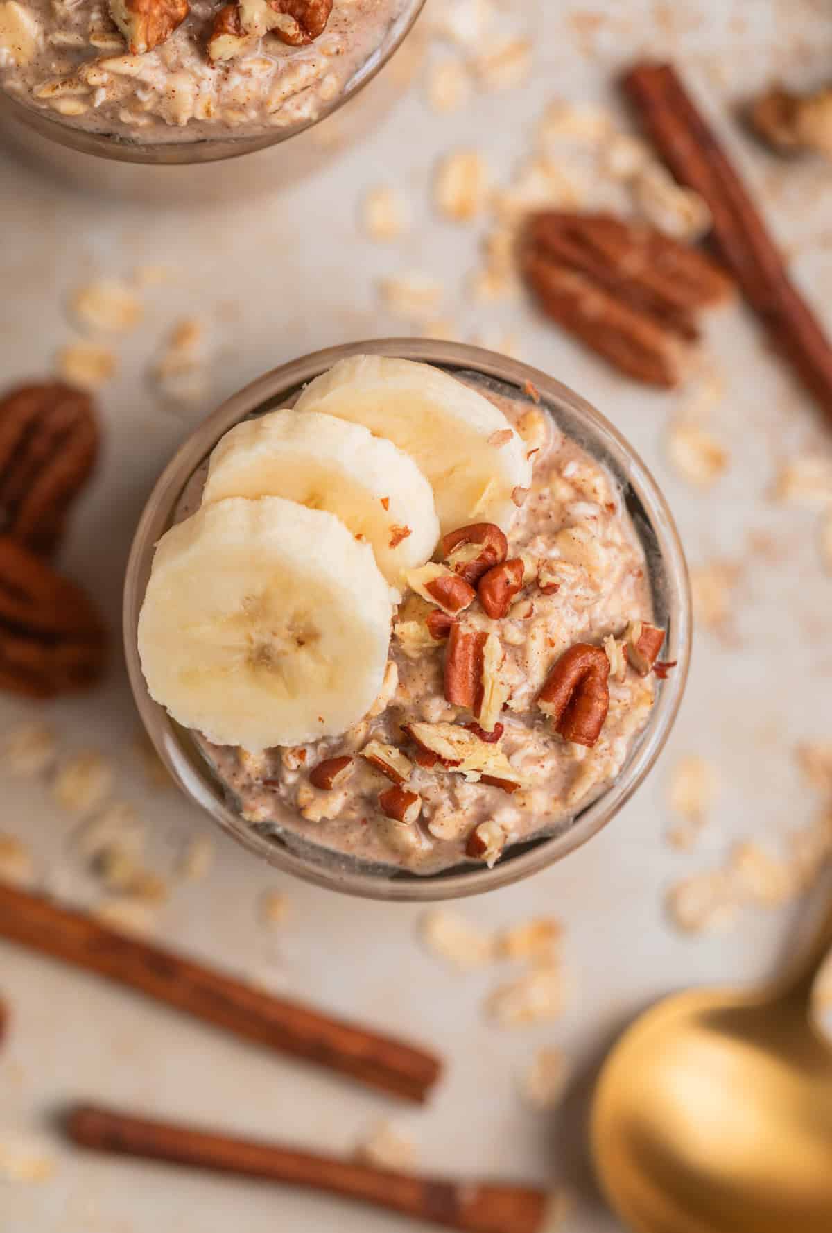 Glass jar with banana bread overnight oatmeal garnished with banana and pecans and cinnamon sticks, pecans and spoon surrounding.