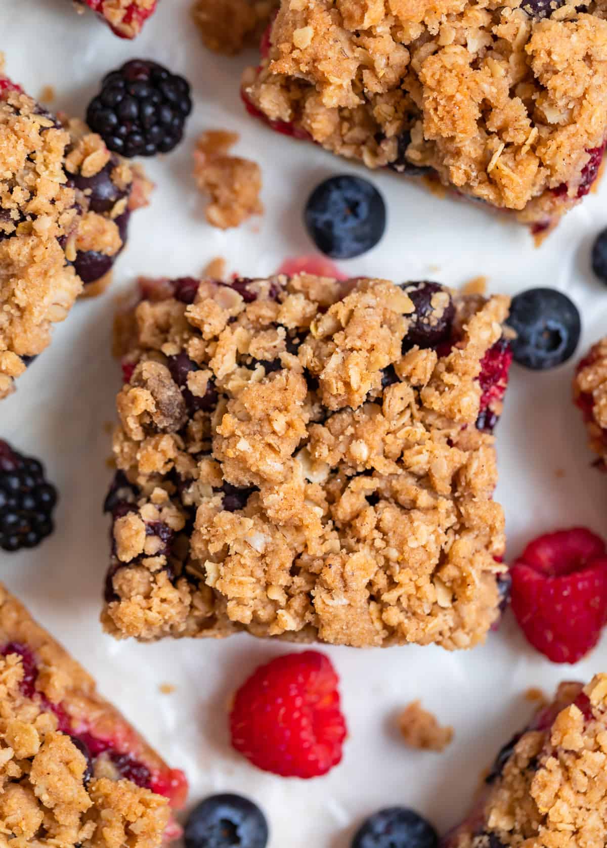 Mixed berry oat bars with crumble topping on white background with fresh berries and crumbles surrounding.