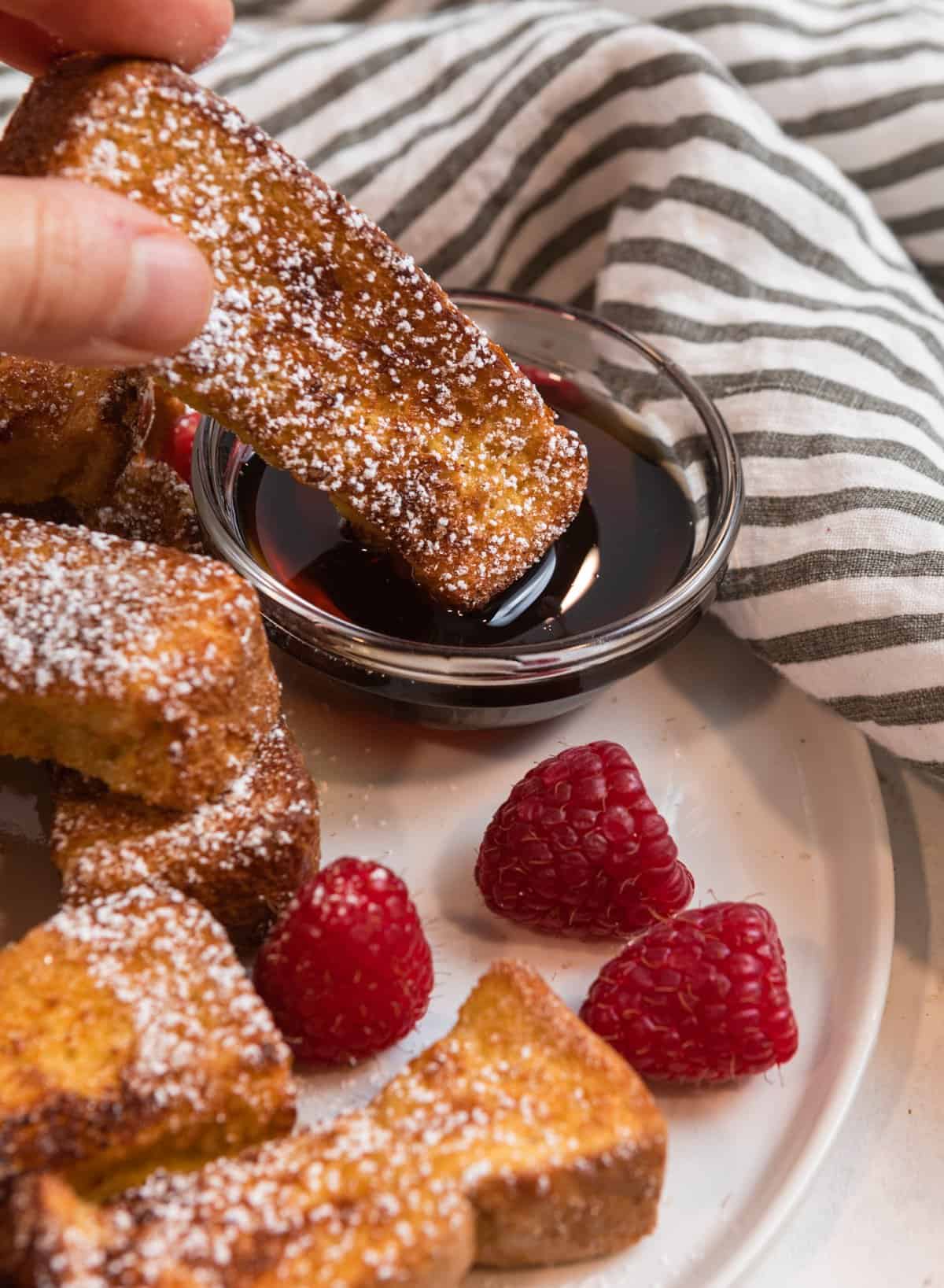 French toast stick being dunked into dish with maple syrup.