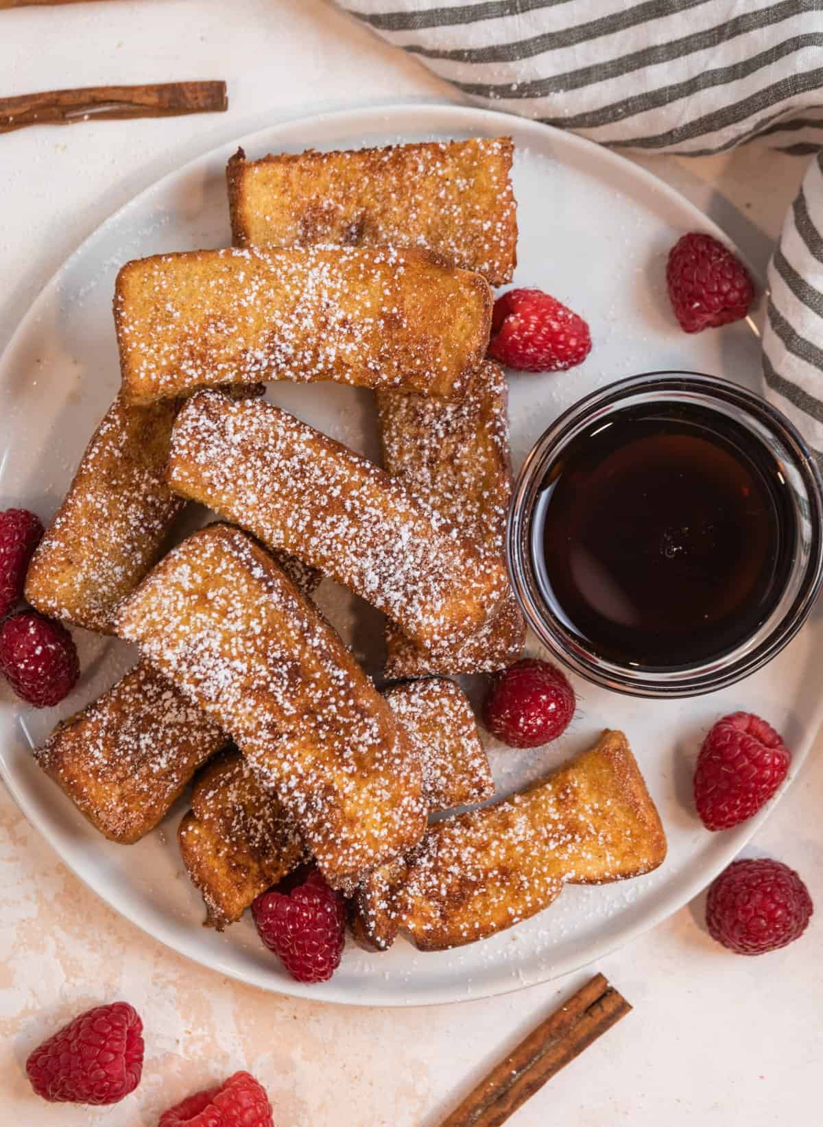 Overhead view of plate of cinnamon sugar covered air fryer French toast sticks with maple syrup in glass bowl.