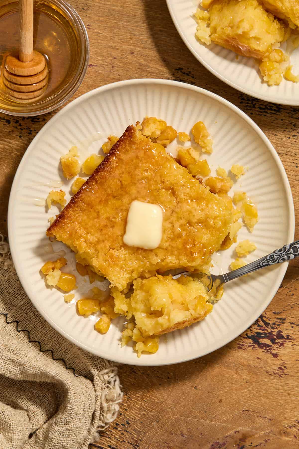 Serving of cornbread casserole on plate with butter and honey on top.