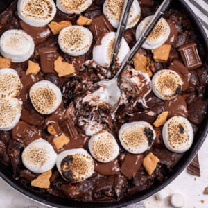 S'mores brownie skillet topped with marshmallows, more chocolate and graham crackers with serving spoons in the center.