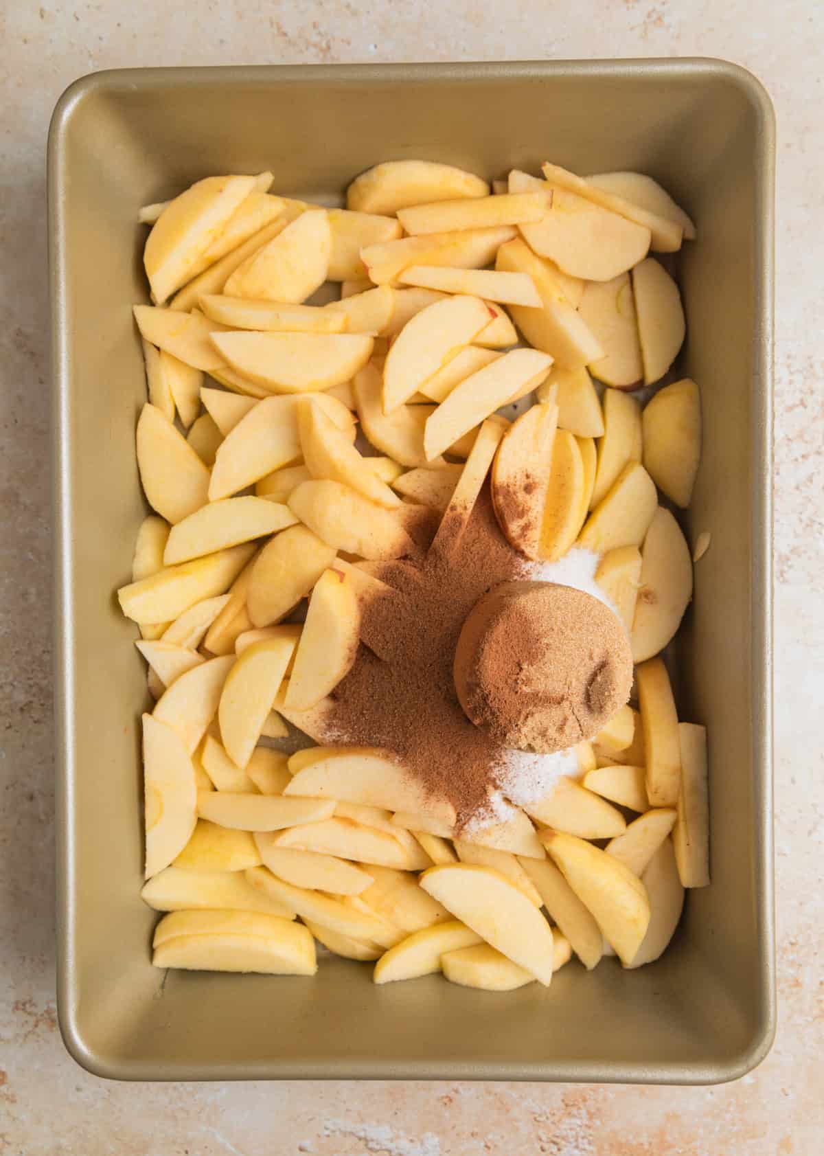 Cinnamon, brown sugar, and white sugar over top of sliced apples in pan.