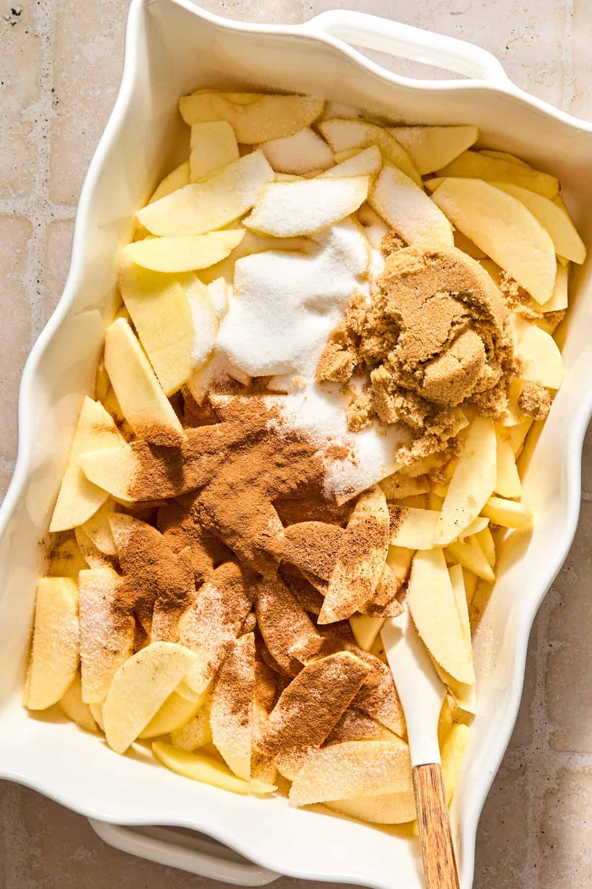 Sliced apples in baking pan with sugar, brown sugar and other ingredients on top.