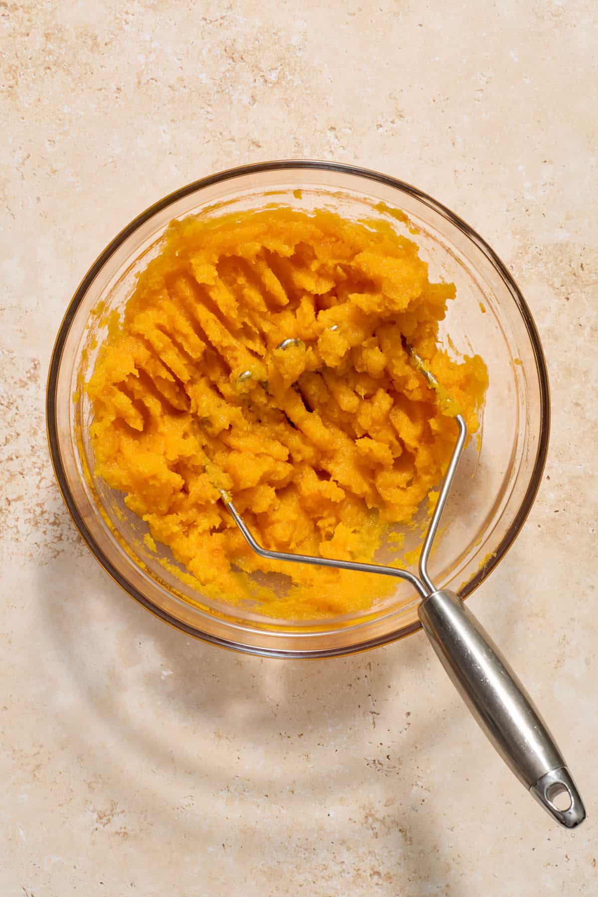 Mashed butternut squash in glass mixing bowl with potato masher.