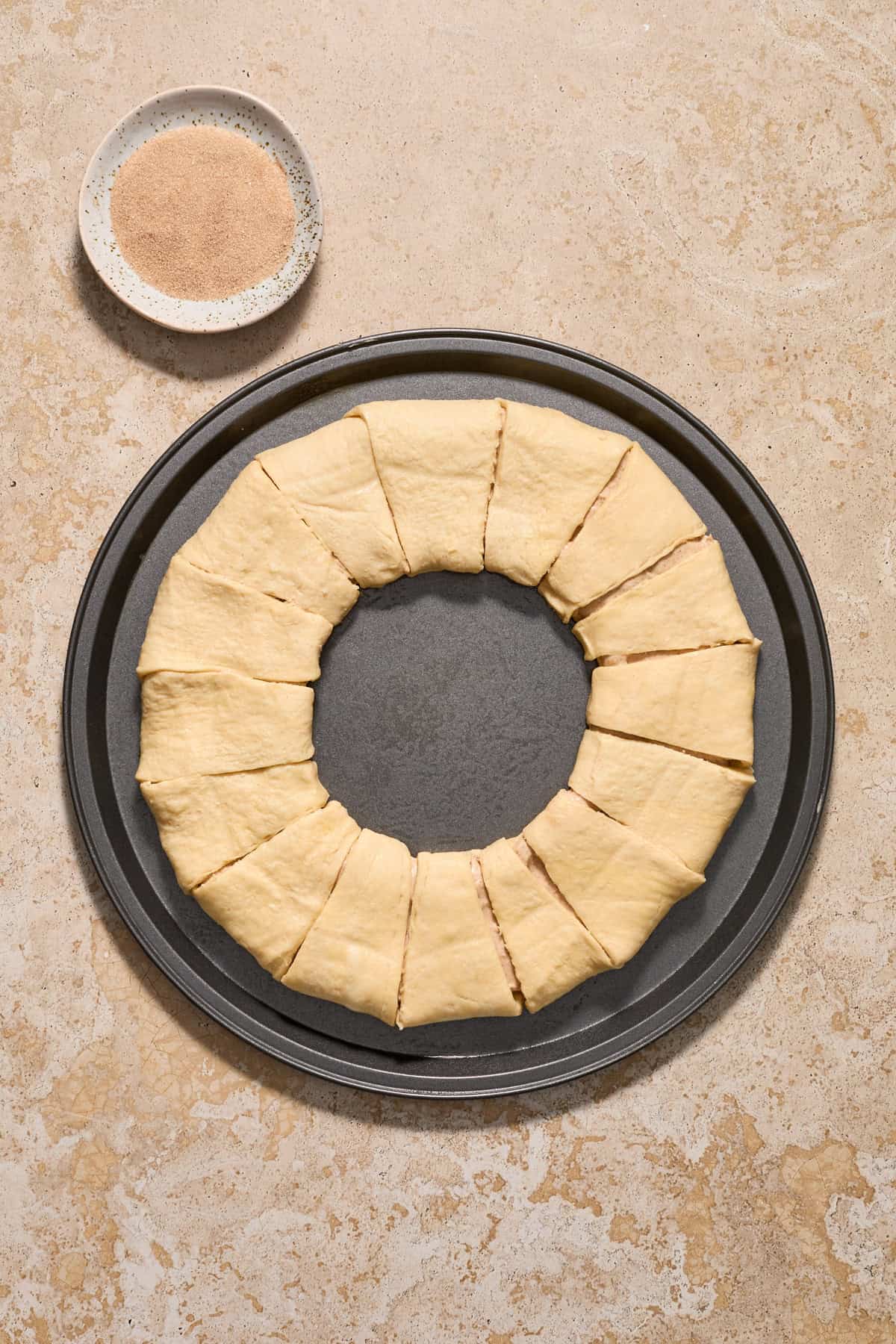 Crescent roll wreath on cookie sheet before baking with cinnamon and sugar in bowl beside it.