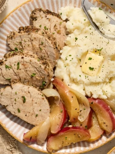 Pork tenderloin sliced on plate with apples and mashed potatoes.