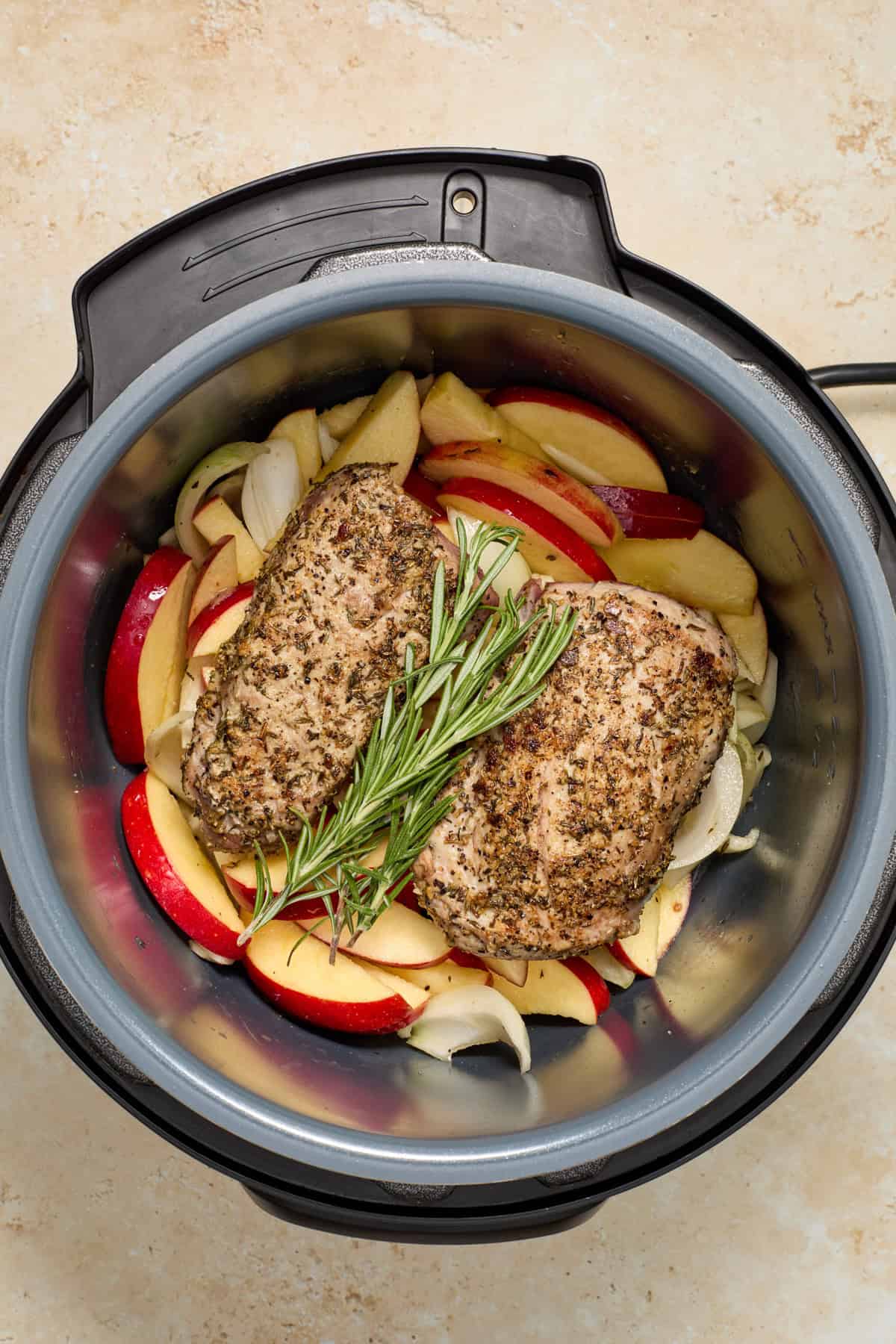 Seared pork tenderloin on top of apples and onions with rosemary sprig in pressure cooker.