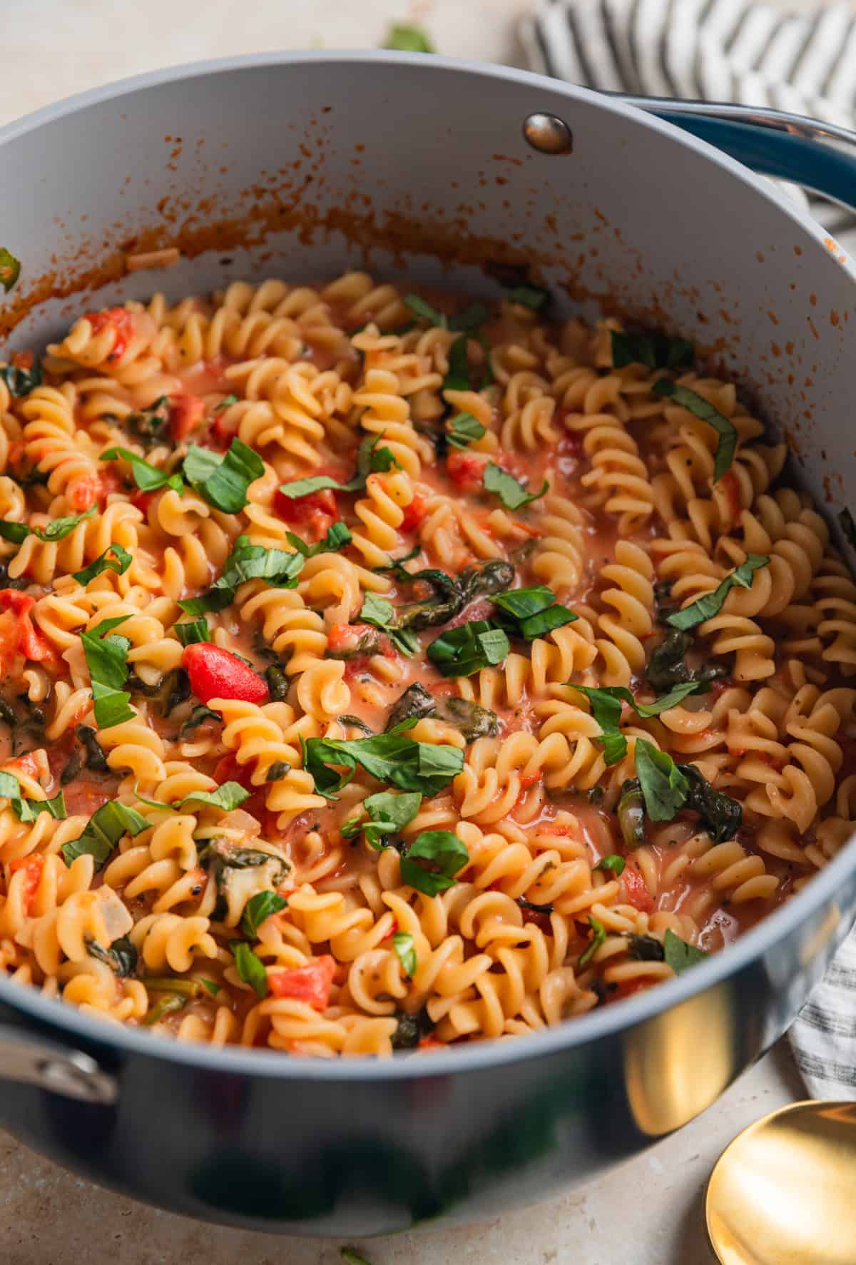 Large pot with creamy rotini pasta, tomatoes, spinach and chopped basil on top.