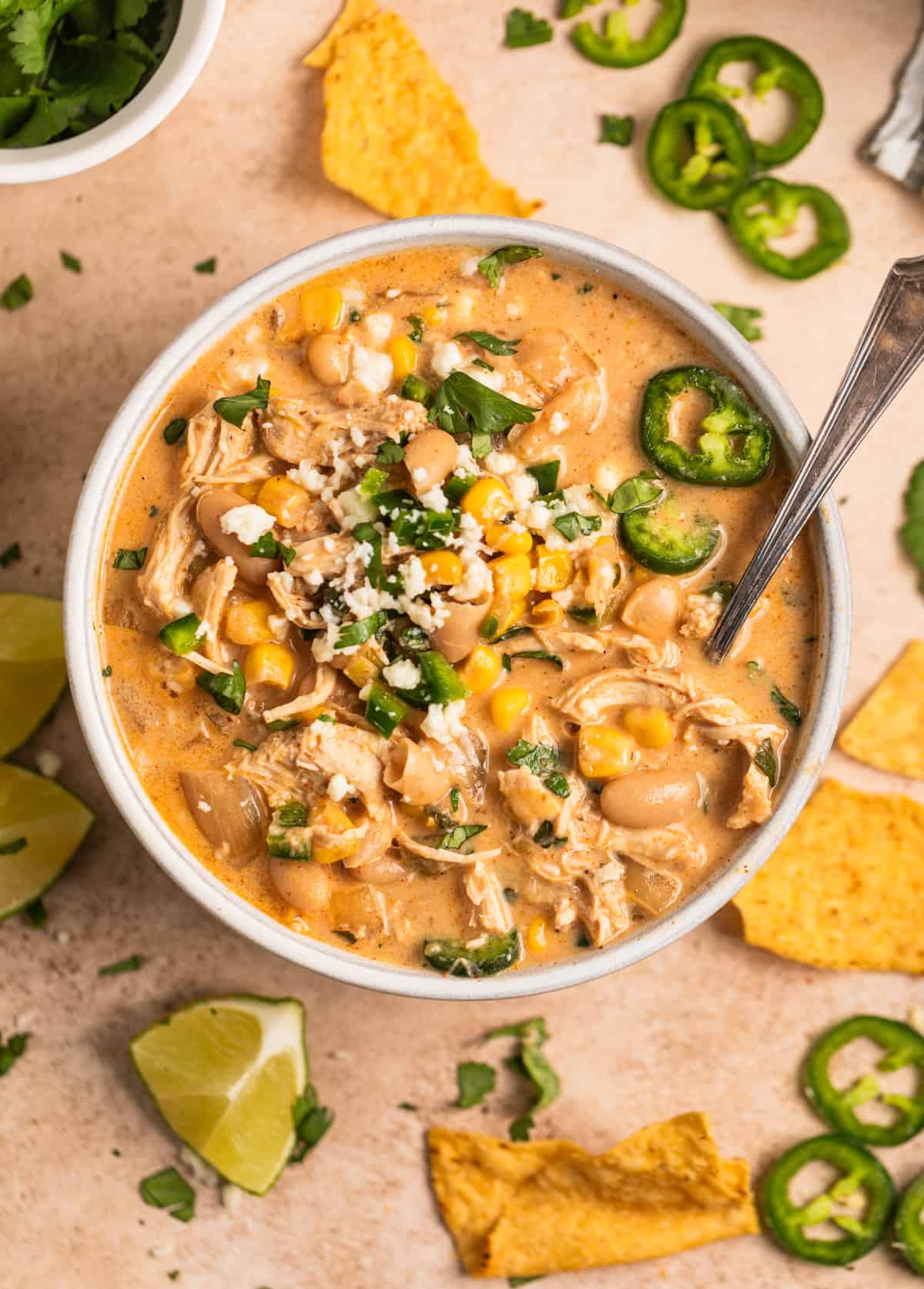 Slow cooker creamy white chicken chili in bowl topped with queso fresca, jalapeno, cilantro and spoon scooping into the soup and chips and limes next to it.