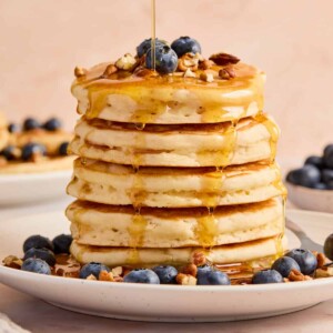 Stack of super fluffy pancakes on plate with blueberries, pecans, and maple syrup.