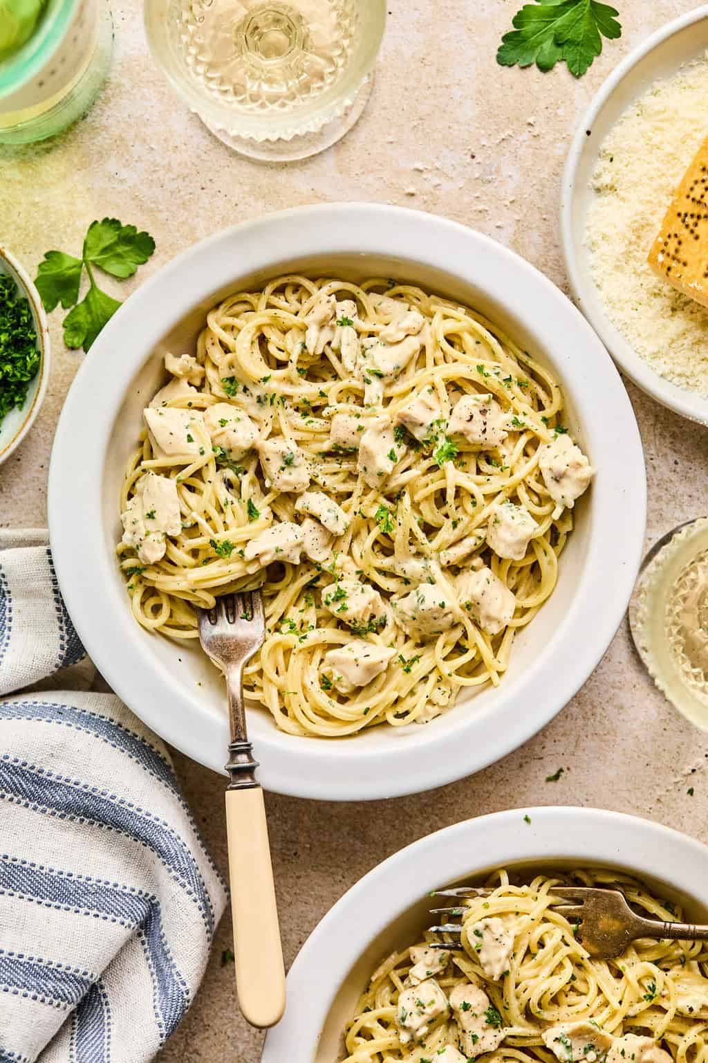 Bowl of chicken Alfredo with napkin, wine, parsley and other ingredients surrounding it.