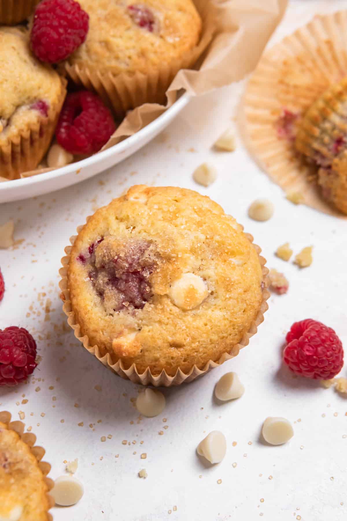 Raspberry white chocolate muffin on counter with morsels and berries surrounding.