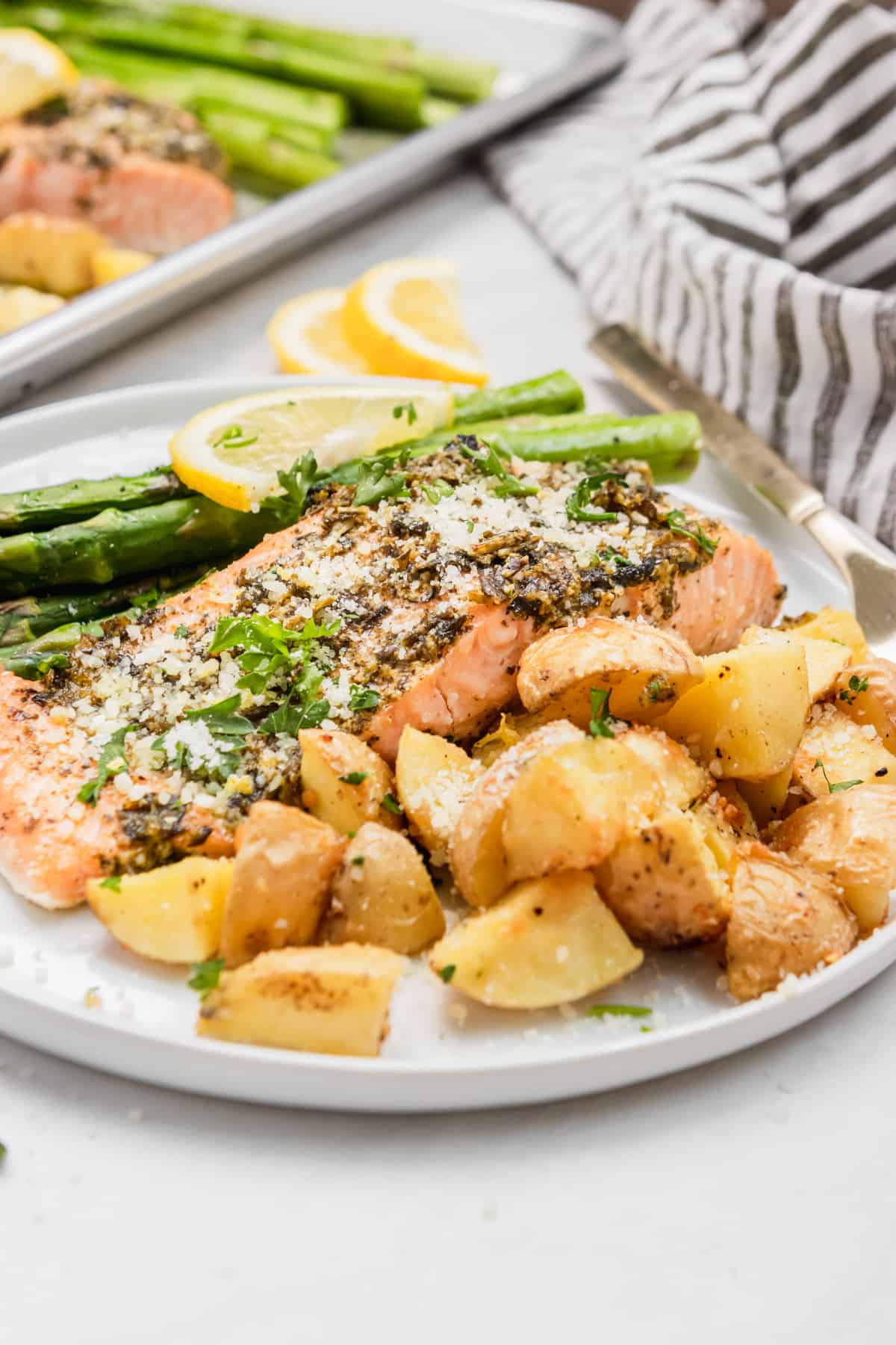 Pesto salmon, potatoes and asparagus on white plate with sheet pan in background.