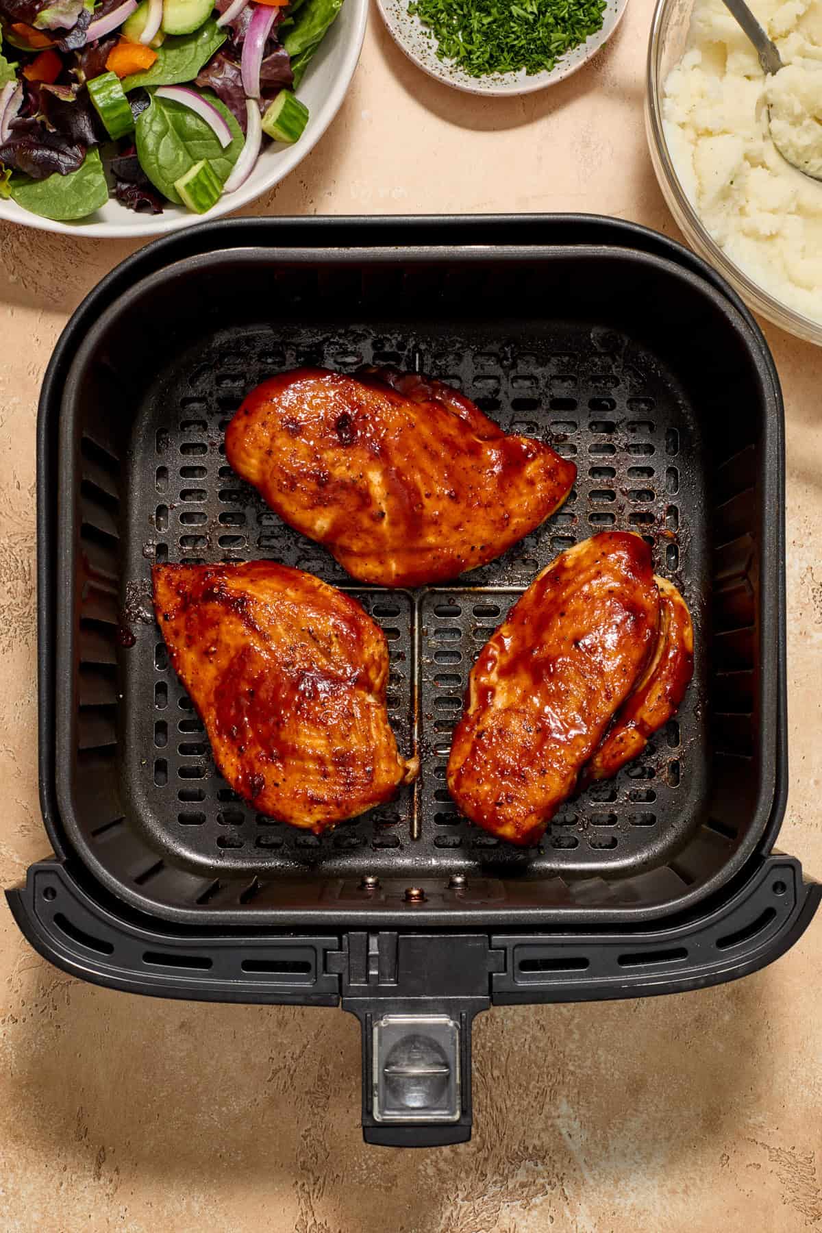 Cooked air fryer bbq chicken breast in air fryer basket with potatoes and salad in bowls beside the air fryer.