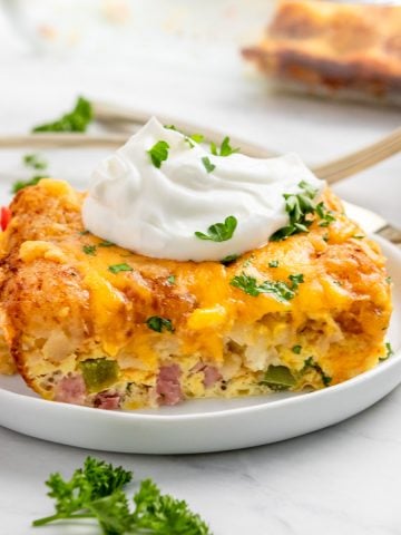 Breakfast casserole with tator tots on white plate with parsley and sour cream.