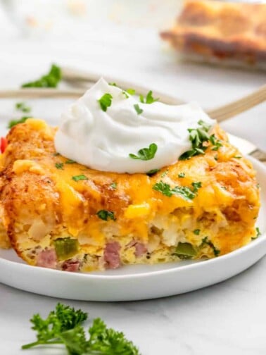 Breakfast casserole with tator tots on white plate with parsley and sour cream.