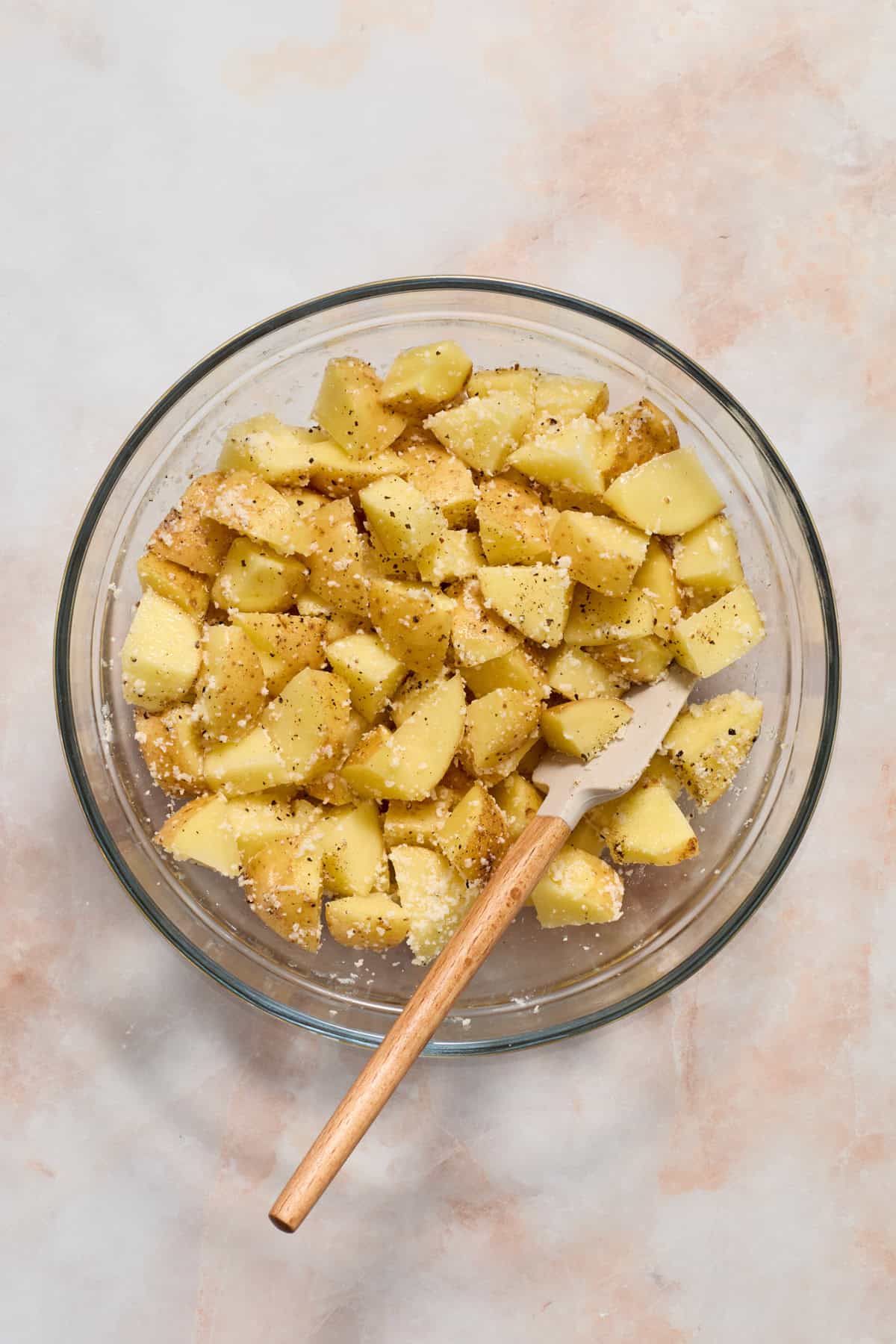 Potatoes cut into bite sized pieces and tossed with parmesan and seasoning in glass bowl with spatula.