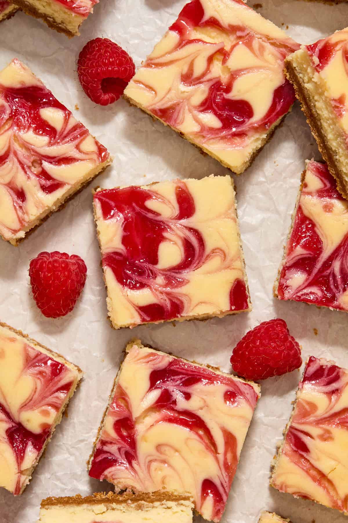 Raspberry swirled cheesecake bars arranged on parchment with raspberries arranged around. 