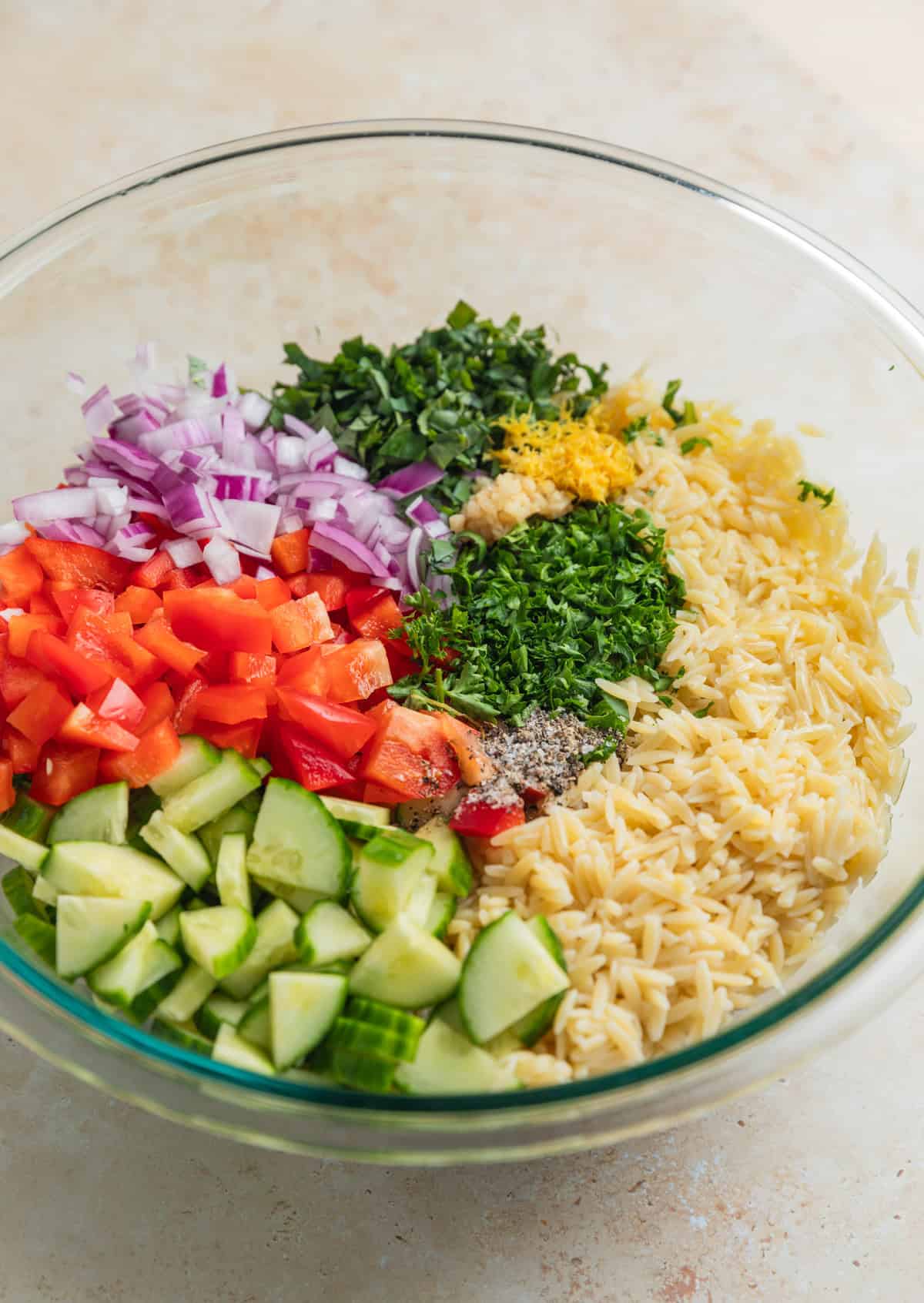 Orzo, lemon zest, cucumber, peppers and other ingredients in glass mixing bowl.