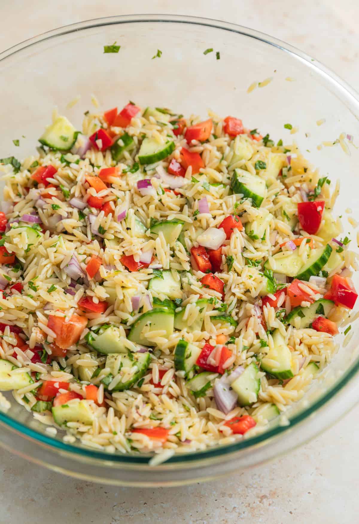 Orzo salad ingredients tossed together in glass mixing bowl before adding feta.