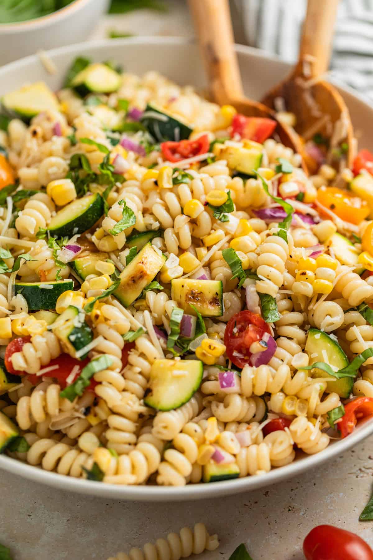 Pasta salad in bowl with grilled zucchini, corn, fresh tomatoes, and topped with basil and parmesan cheese.