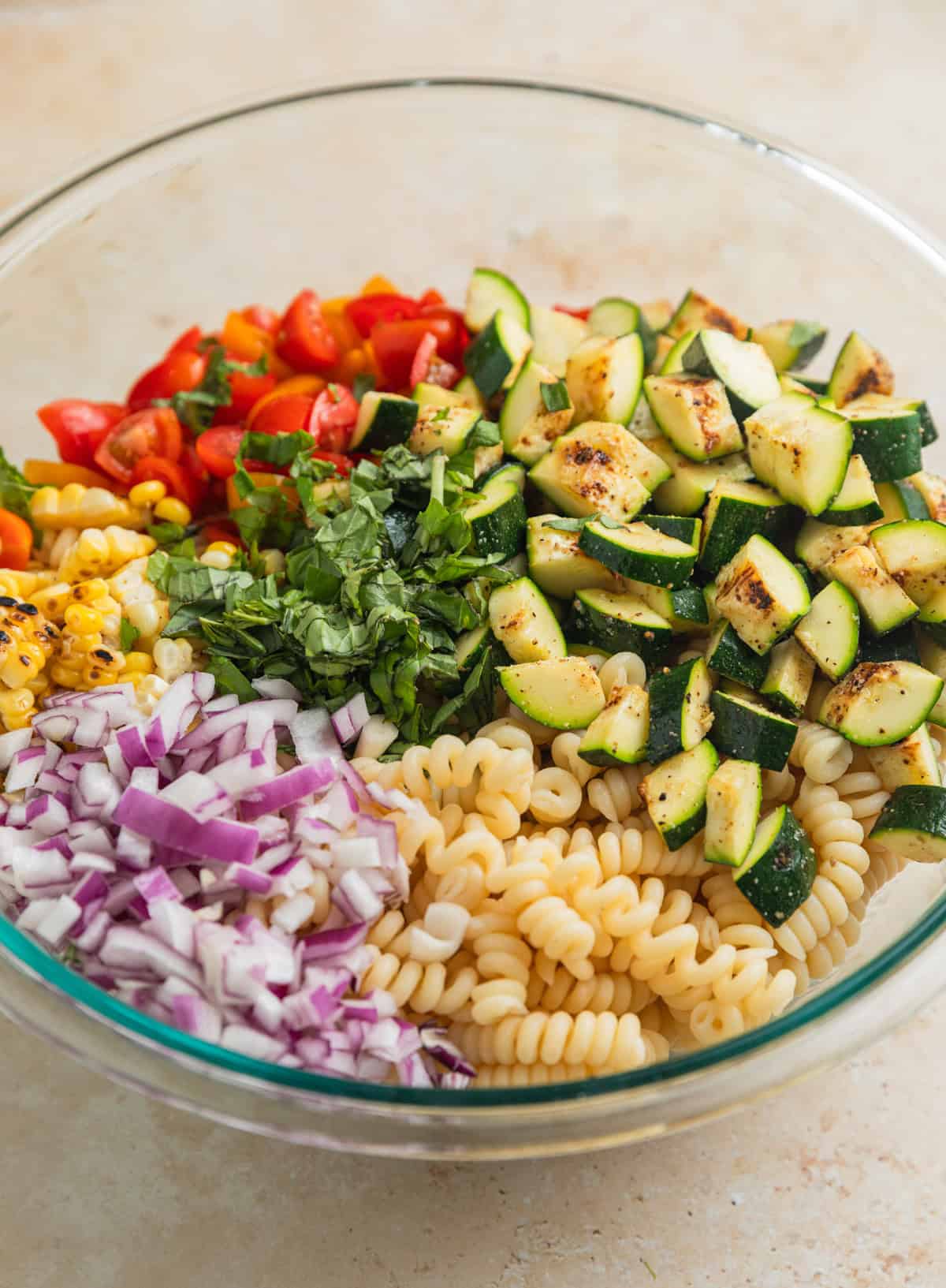 Cooked pasta, red onion, tomatoes, corn and zucchini in glass mixing bowl.
