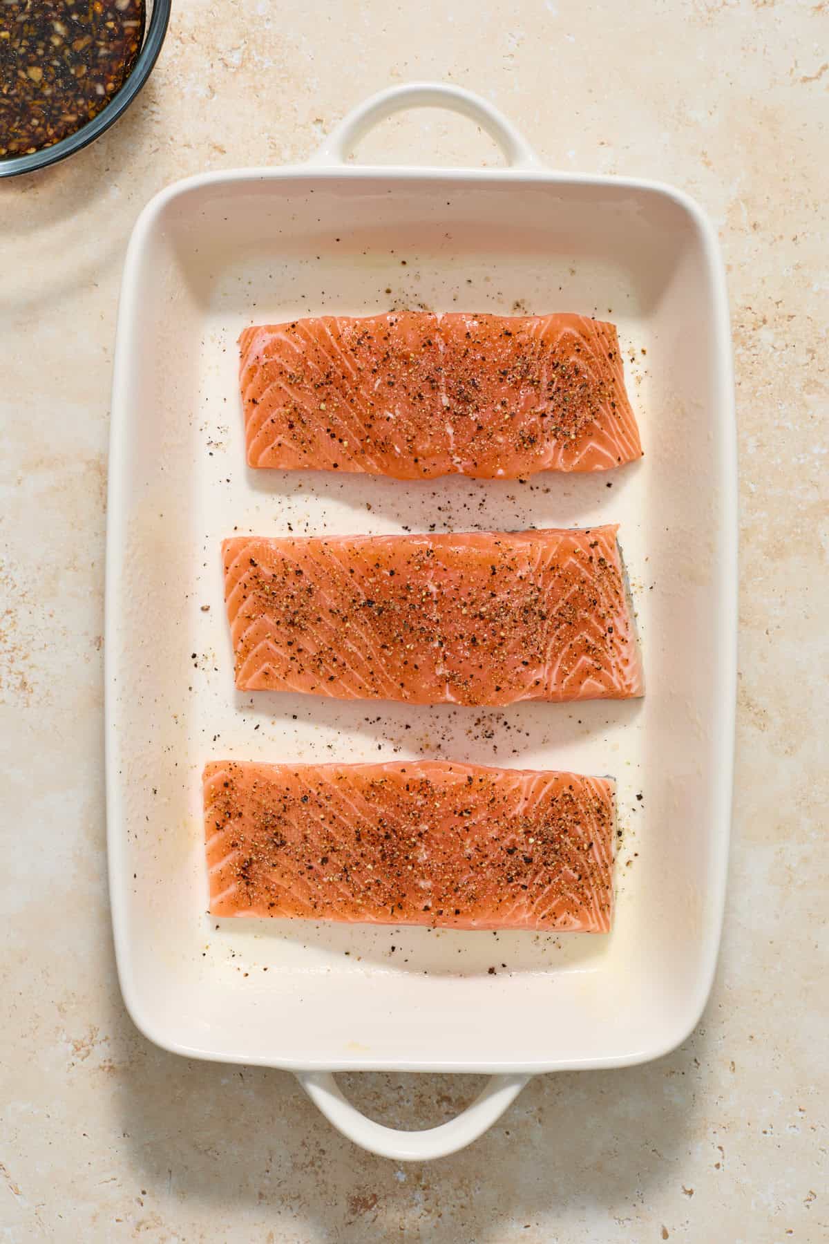Salmon fillets in white baking pan seasoned with salt and pepper.