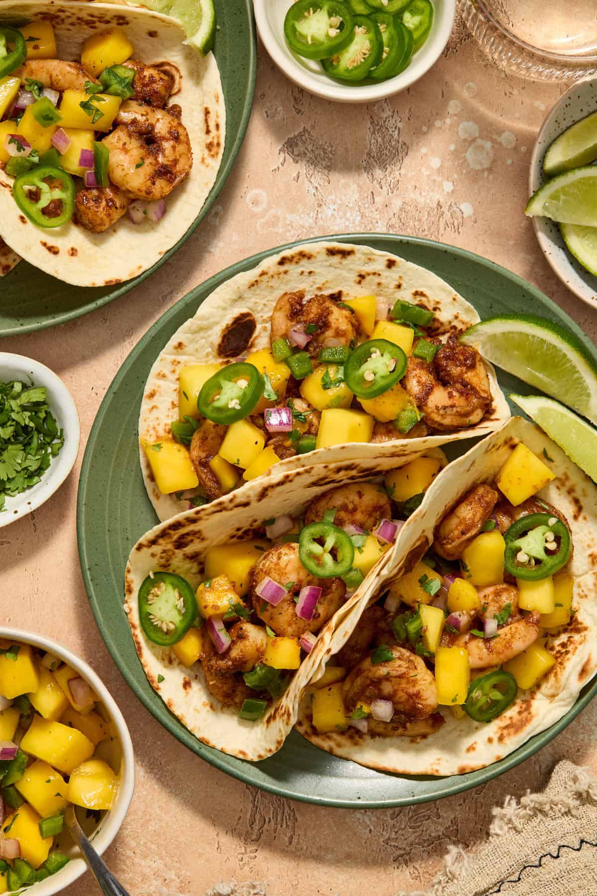 Spicy shrimp tacos on green plate topped with mango salsa, sliced jalapeño and lime wedges on the side.