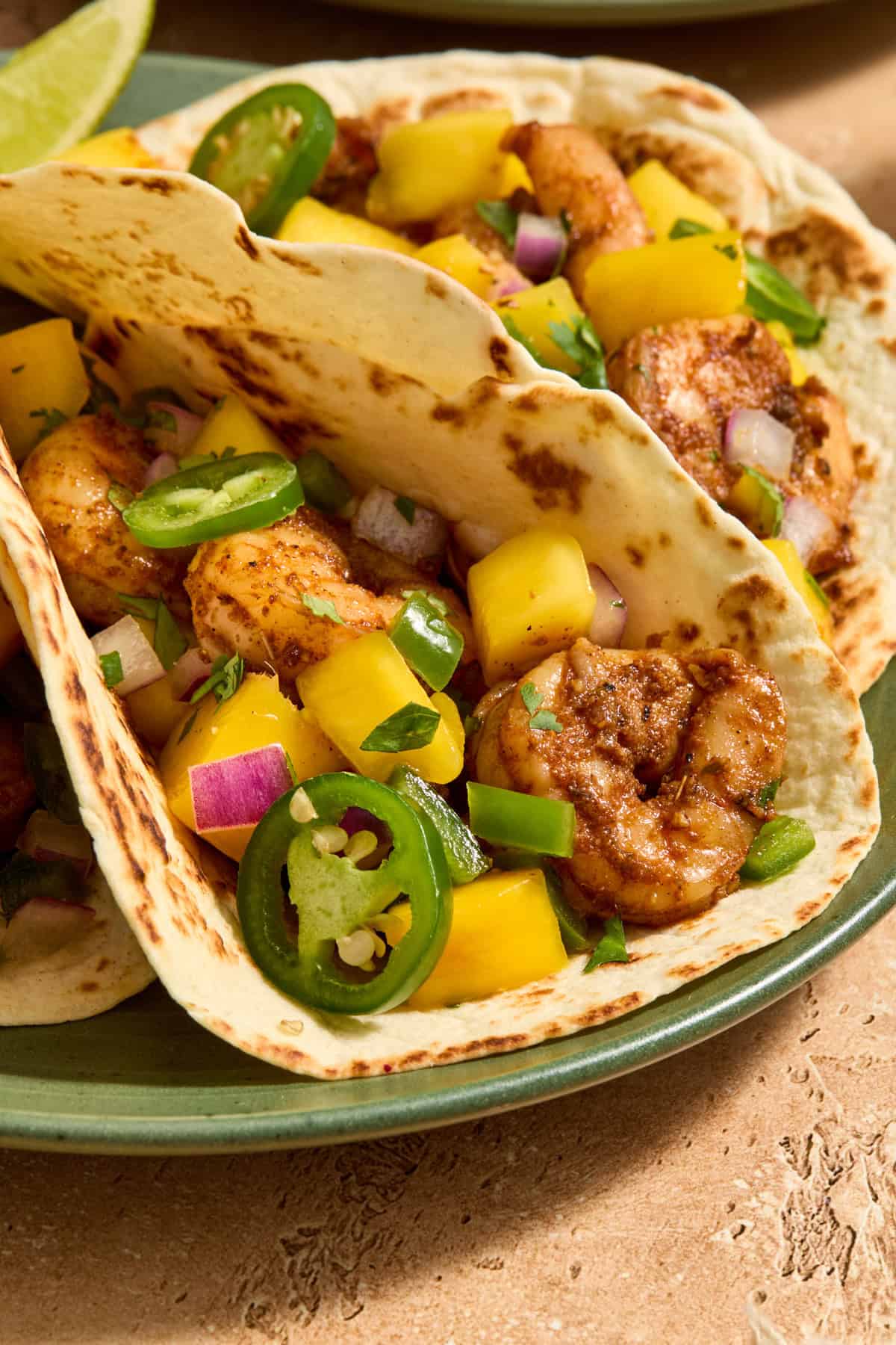 Spicy shrimp tacos on plate topped with mango salsa, cilantro and sliced jalapeños.