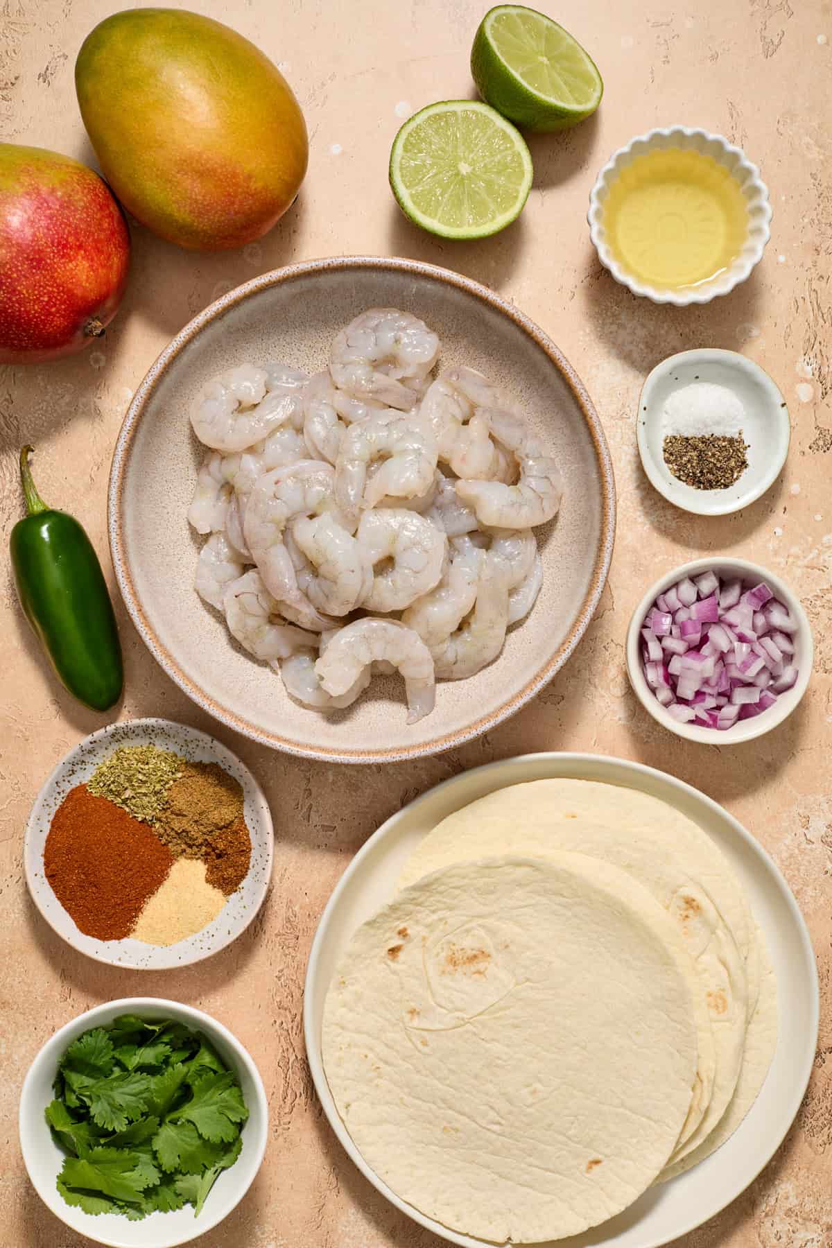 Raw shrimp, mango, spices, tortillas, limes and other ingredients arranged on surface.