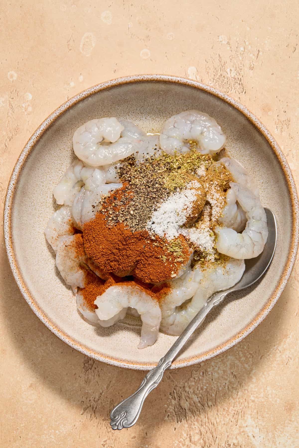 Chili powder, garlic powder and other seasoning on raw shrimp in bowl with spoon.