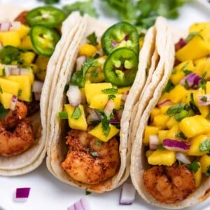 Spicy shrimp tacos on white plate topped with mango salsa and slices of jalepeño.