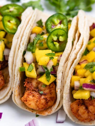 Spicy shrimp tacos on white plate topped with mango salsa and slices of jalepeño.