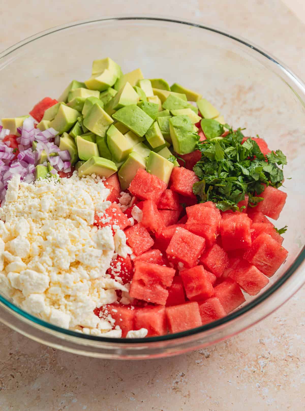 Diced avocado, feta cheese crumbles, cilantro, onion and watermelon cubes in glass bowl before mixing.