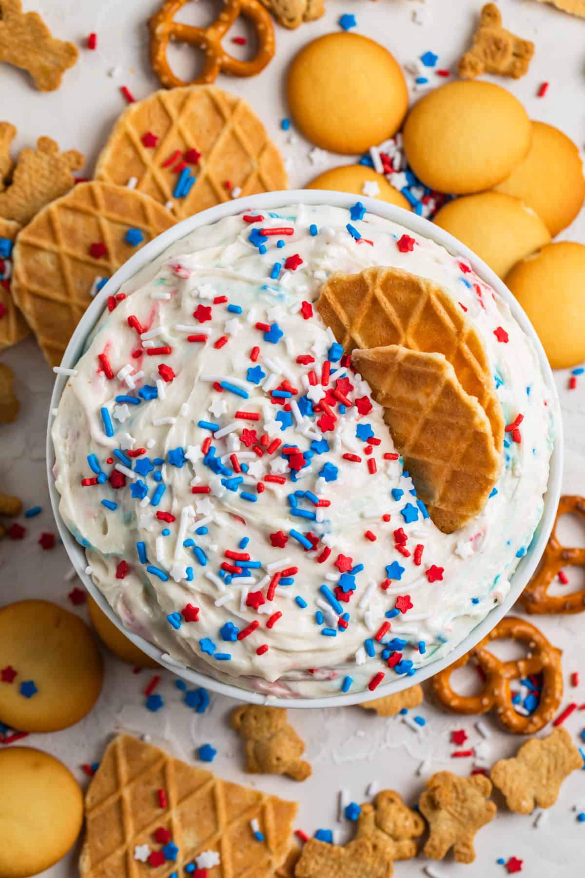 Dunkaroo dip in white bowl with red, white and blue sprinkles on top and cookies and pretzels surrounding.