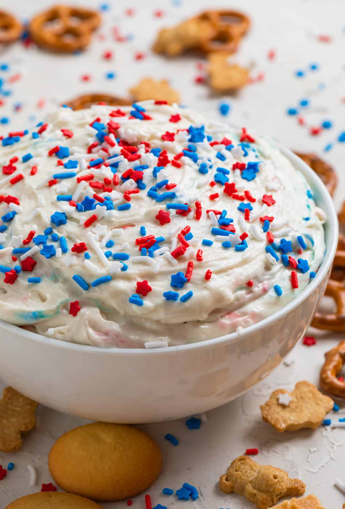 Red, white and blue patriotic funfetti dip in white bowl with sprinkles on top and cookies surrounding.