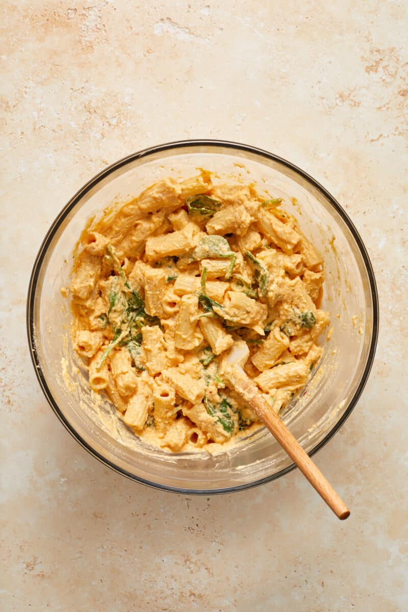 Pumpkin mixture tossed with pasta and spinach in glass mixing bowl with spatula.