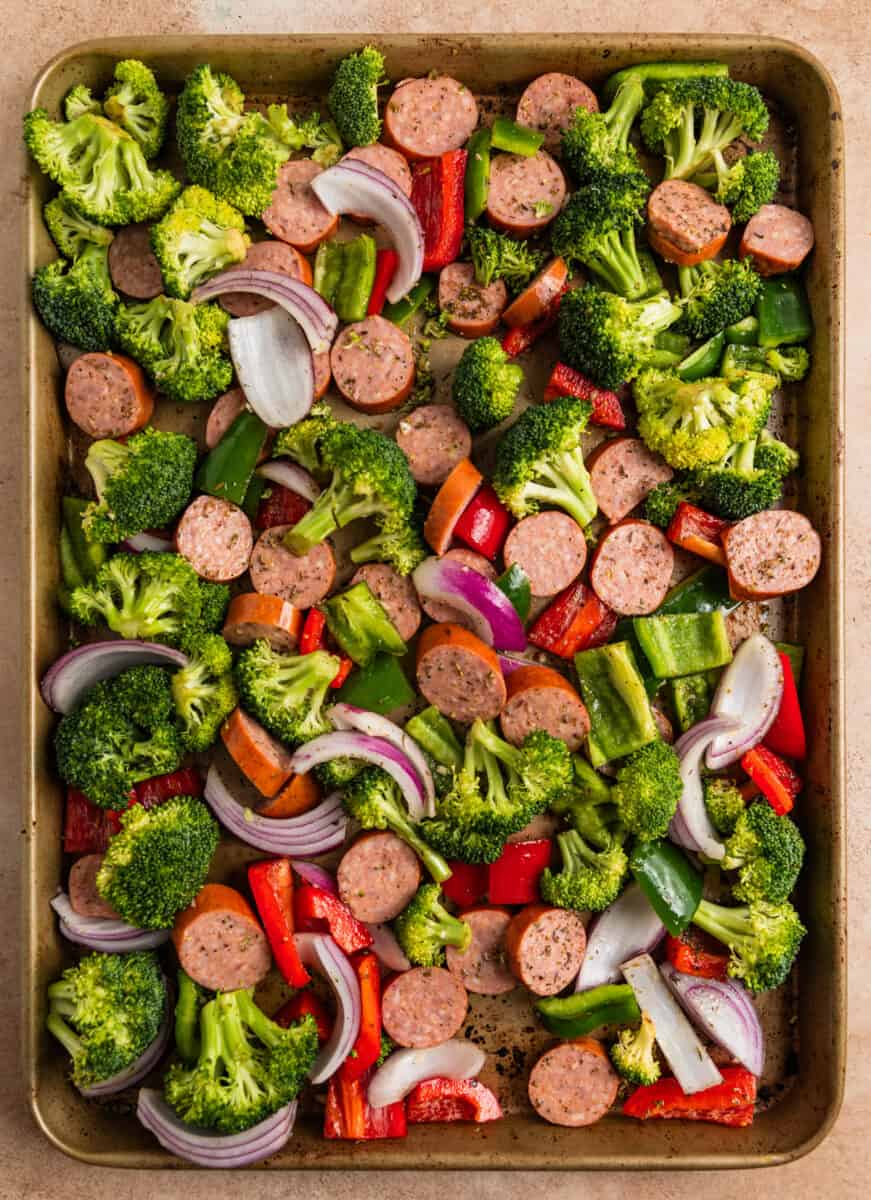 Sausages and veggies tossed in olive oil and seasoning on baking sheet.