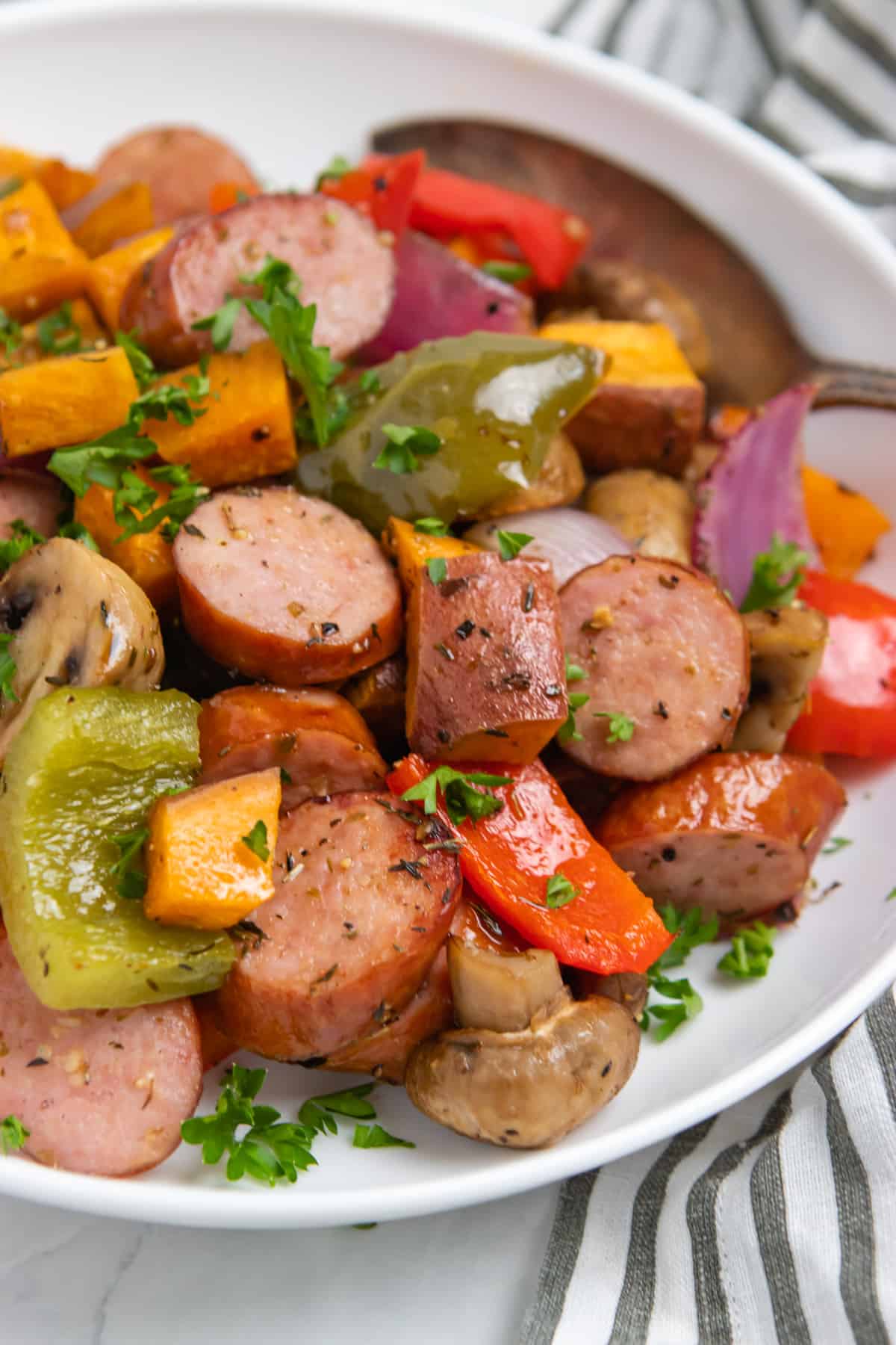 Bowl of cooked sausages, peppers and other veggies with spoon.