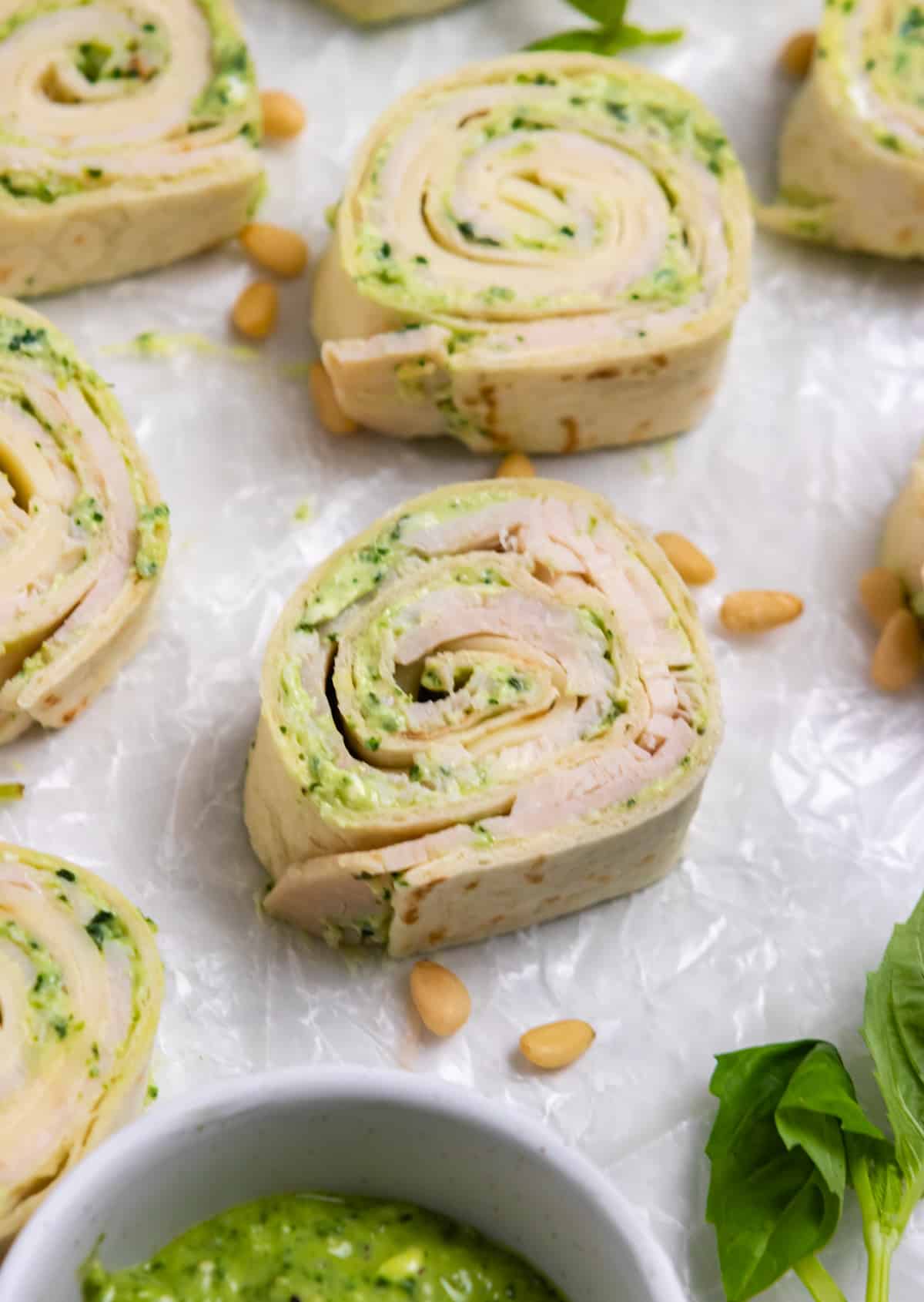 Turkey pinwheels with pesto on wax paper with basil and pine nuts surrounding.