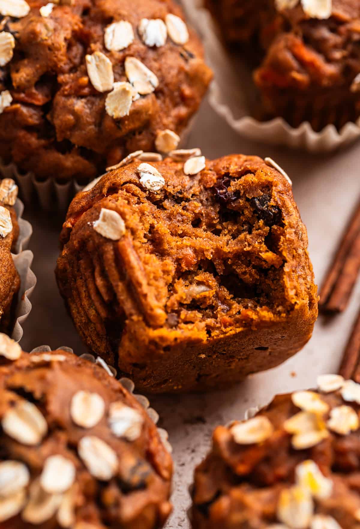 Bite shot of pumpkin carrot muffin surrounded by other muffins and two cinnamon sticks.