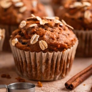 Pumpkin muffins with carrots topped with rolled oats on counter with cinnamon sticks and measuring spoons beside them.