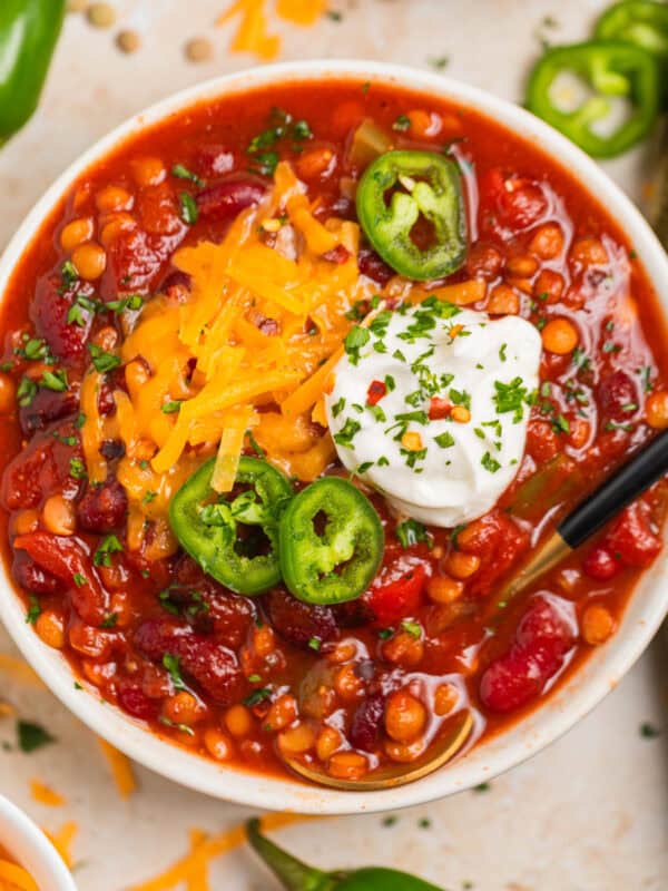 Lentil chili in bowl with beans and peppers and topped with cheese and other toppings with spoon scooping into the bowl.