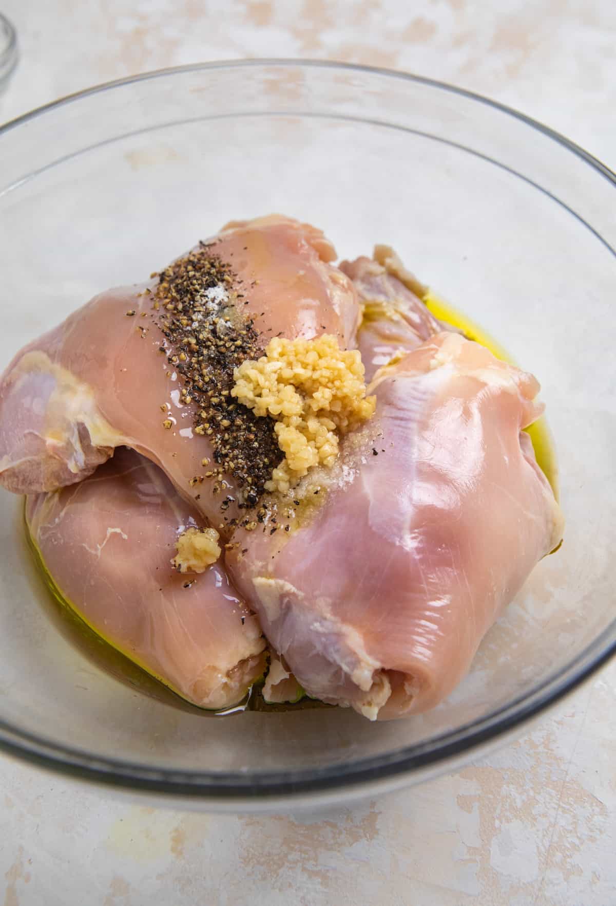 Mixing bowl with raw chicken thighs, olive oil, garlic and seasoning.