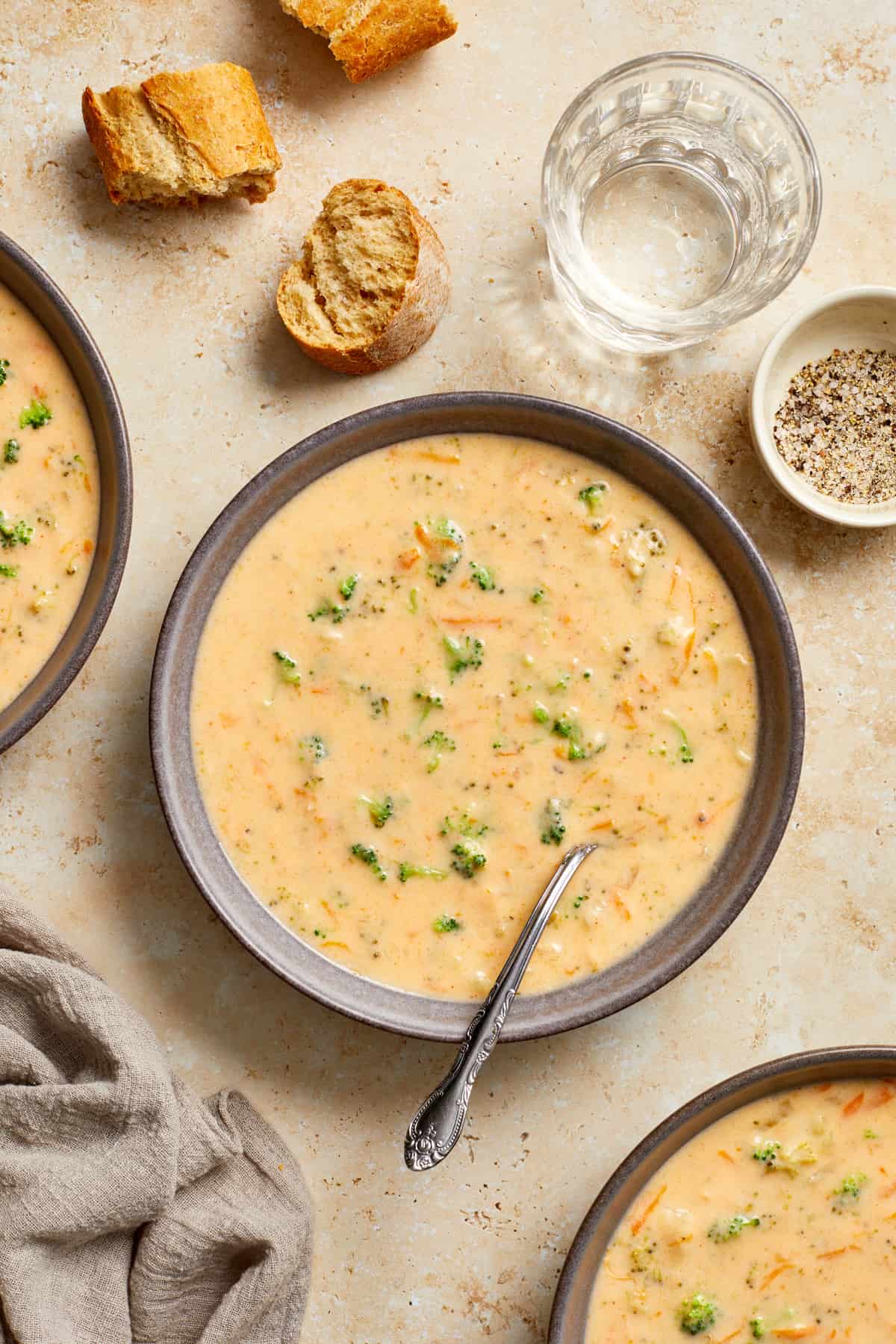 Bowl of broccoli and cheese soup with spoon surrounded by glass with water, pieces of bread and other bowls of soup.